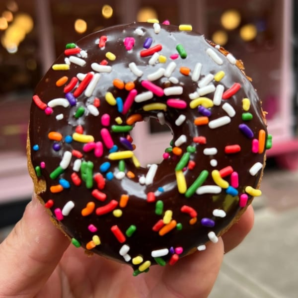 A hand holds a Dat Donut with chocolate frosting and colorful sprinkles, against a blurred background.