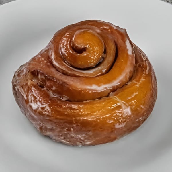 A glazed cinnamon roll from Dat Donut, with a shiny, sweet coating on a white plate.