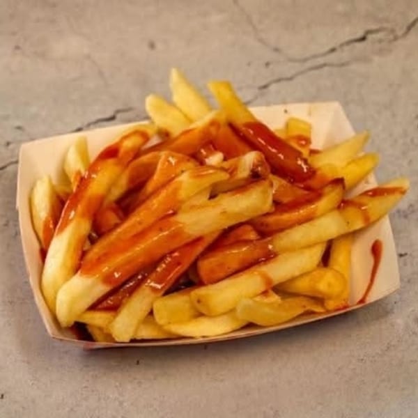A cardboard tray filled with golden french fries, drizzled generously with red ketchup, on a gray surface.